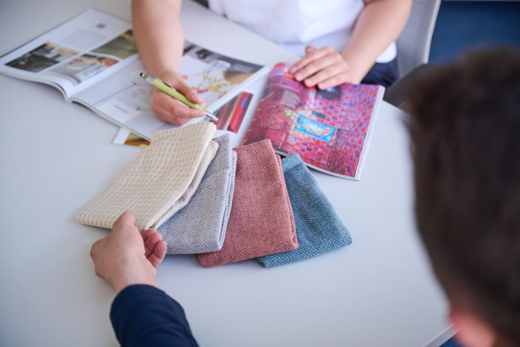 Franziska Schwartz zeigt einem Kunden verschiedene Stoffe in beige, blau und Beere, ebenso liegt ein Interior Magazin auf dem Tisch und der Kunde fasst ein Beiges Material an