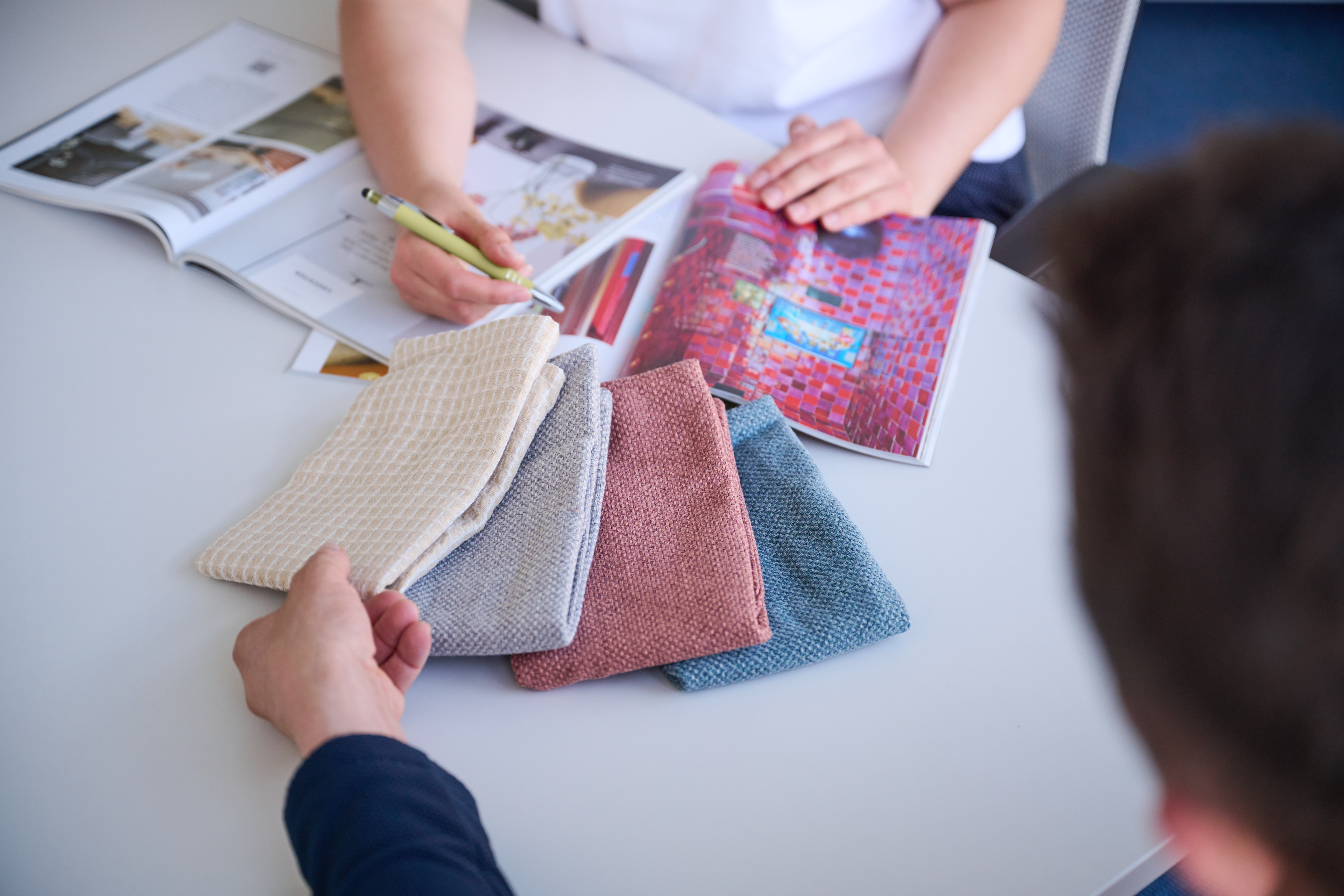 Franziska Schwartz zeigt einem Kunden verschiedene Stoffe in beige, blau und Beere, ebenso liegt ein Interior Magazin auf dem Tisch und der Kunde fasst ein Beiges Material an