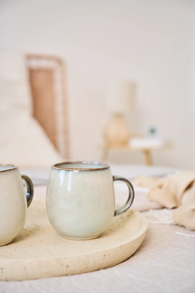 Detailansicht von Deko, 2 Steingut Tassen im Vordergrund und ein Nachttisch in hellen Farben mit Naturmaterialien im Hintergrund