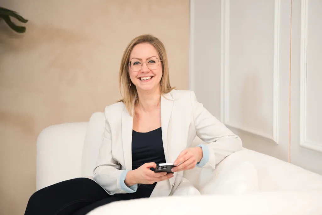 Franziska Schwartz sitzend auf Couch heller Hintergrund, schaut vom handy in Richtung des Betrachters